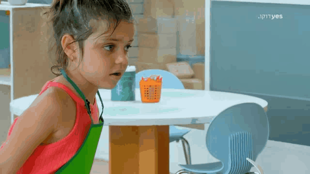 a little girl wearing an apron is standing in front of a table with a basket of pencils on it .