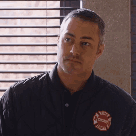 a man wearing a chicago fire department jacket is standing in front of a window with blinds .