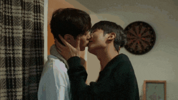 two young men are kissing in a room with a dart board in the background .
