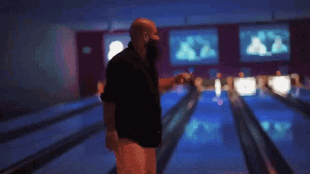 a bald man with a beard is standing in a bowling alley holding a cell phone