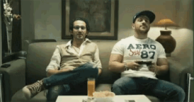 two men are sitting on a couch playing a video game and one of them is wearing an aero 87 shirt