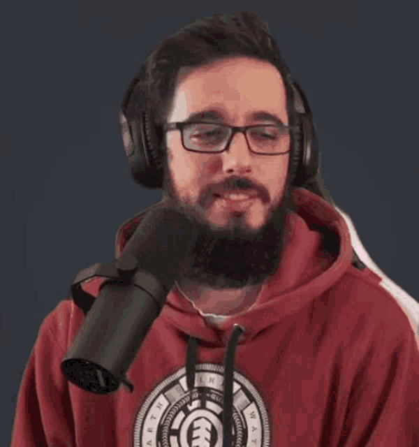 a man with a beard wearing headphones and a red hoodie is smiling in front of a microphone