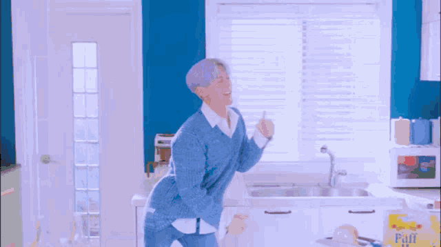 a man in a blue sweater is dancing in a kitchen with a bag of puff corn in the background .