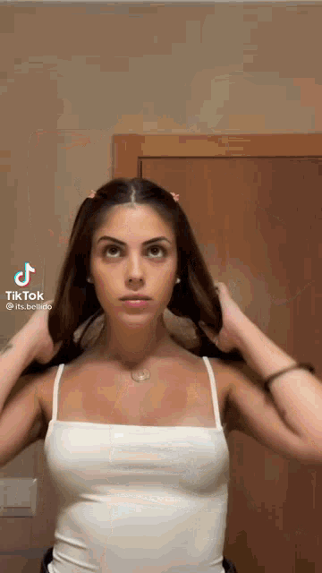 a woman wearing a white tank top is holding her hair in front of a door .