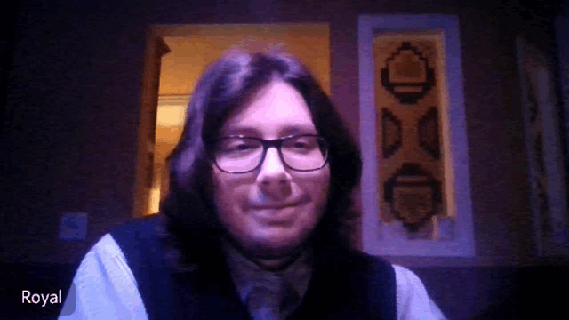 a man with long hair and glasses is smiling in front of a dark background and the name royal is on the bottom right
