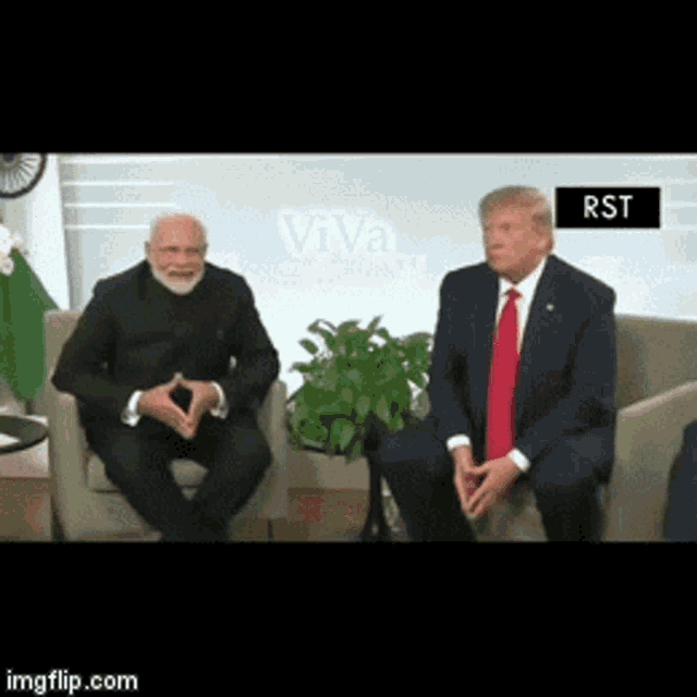 a man in a suit and tie sits next to another man in a suit and tie in front of a sign that says rst