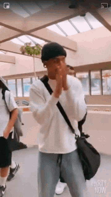 a man wearing a white sweatshirt and a black beanie is standing in a hallway with his hands folded .