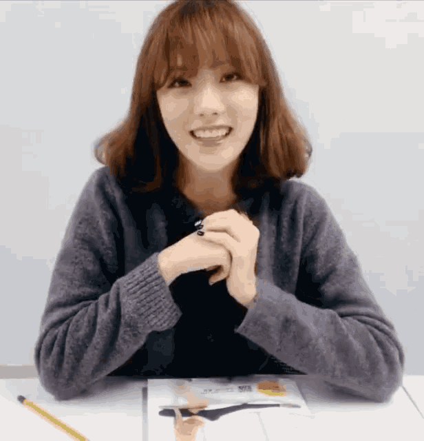a woman in a grey sweater sits at a table with her hands folded in front of her face