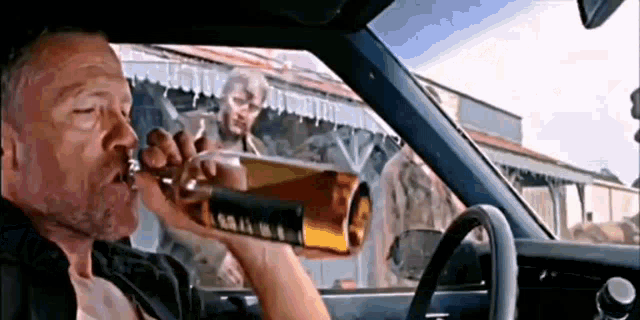 a man is driving a car and drinking from a bottle of whiskey .