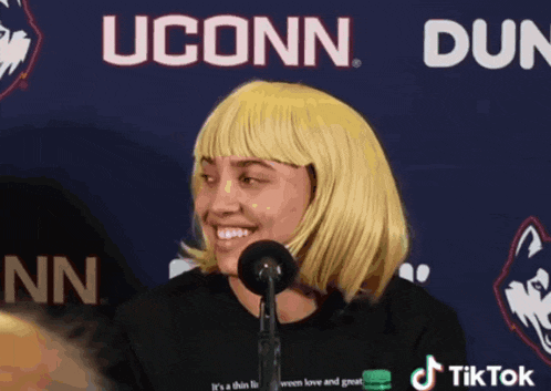 a woman wearing a blonde wig is smiling in front of a uconn logo