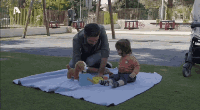 a man is playing with a baby on a blanket in a park with the letter a in the background