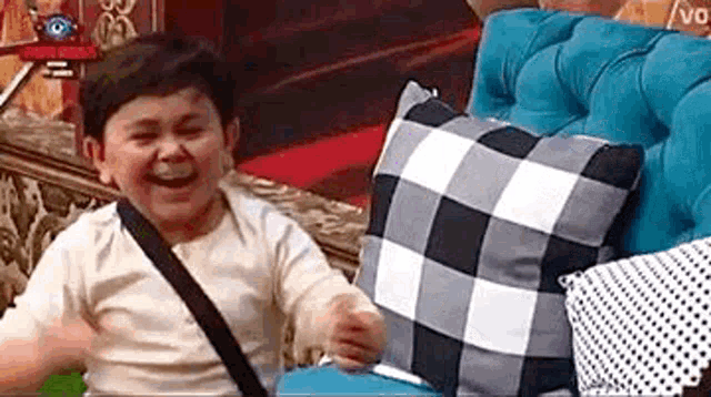 a little boy is sitting on a blue couch with pillows and laughing .