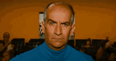 a bald man wearing a blue turtleneck sweater looks at the camera with a serious look on his face .