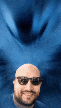 a bald man with a beard wearing sunglasses is smiling in front of a blue background