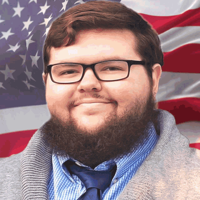 a man with glasses and a beard is smiling in front of a flag