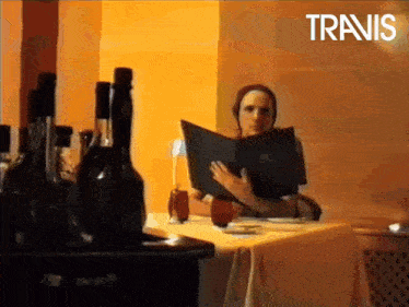 a woman sits at a table reading a menu with the word travis in the upper left corner