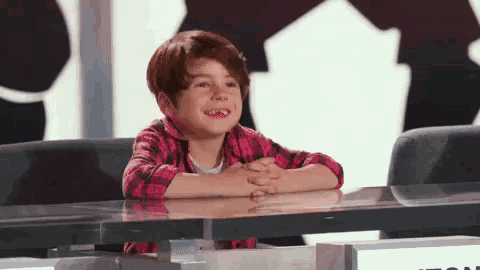 a young boy in a plaid shirt is sitting at a table with his hands folded