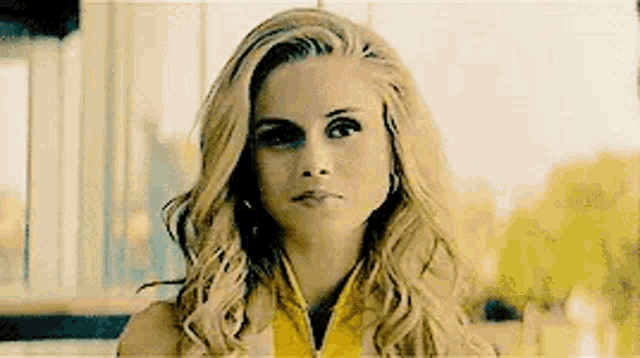 a close up of a woman with long blonde hair wearing a yellow jacket and earrings .