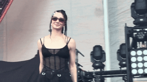 a woman wearing sunglasses and a black tank top stands in front of a row of lights