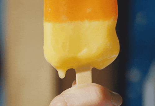 a person is holding an orange and yellow popsicle on a wooden stick