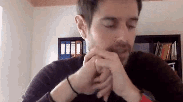 a man is sitting in front of a bookshelf with his hands folded in prayer .