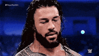 roman reigns is a wrestler with long hair and a beard and is looking down .