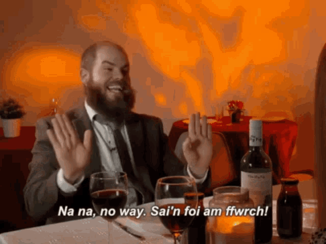 a man with a beard is sitting at a table with wine glasses and a bottle of wine and says na na no way