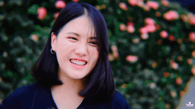 a girl with purple hair is smiling in front of a bush with pink flowers