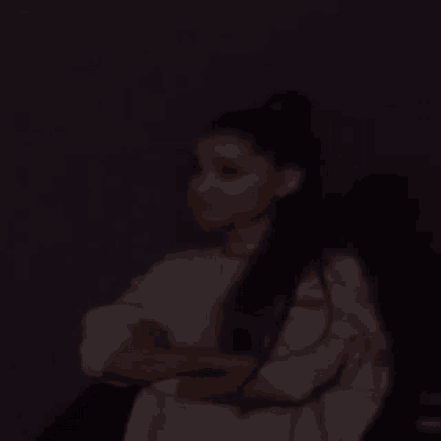a woman in a white shirt is sitting in a dark room with her arms crossed .
