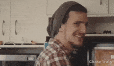 a man wearing a beanie and plaid shirt is smiling in front of a microwave oven .