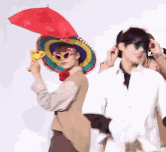 a man wearing a sombrero and sunglasses is holding a red umbrella while standing next to another man .