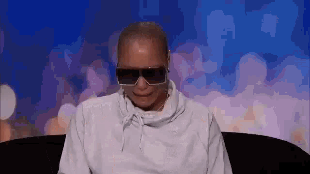 a bald man wearing sunglasses and a white hoodie is sitting on a couch .