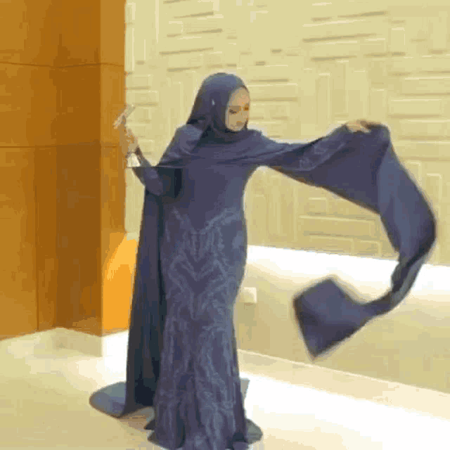 a woman in a blue dress and hijab is standing in front of a wall holding a scarf .