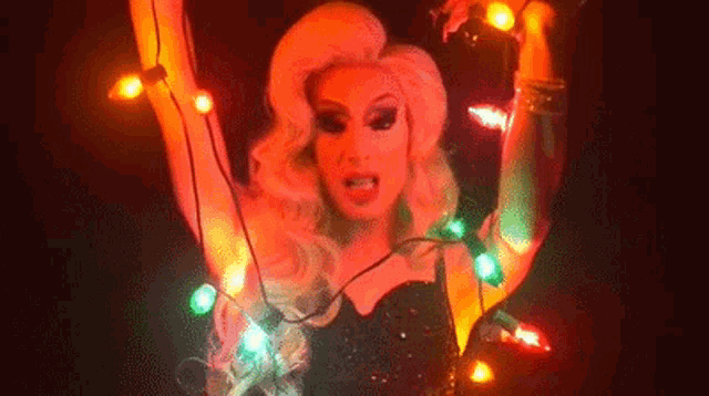 a woman in a drag queen costume is holding a string of christmas lights .