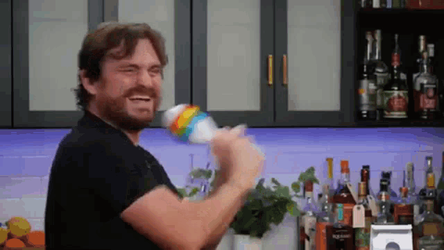 a man in a black shirt is holding a rainbow colored can in front of a bar filled with bottles of alcohol .