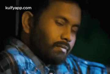 a man with a beard is smoking a cigarette while wearing a blue plaid shirt .