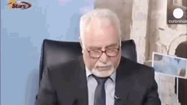 an older man with a beard and glasses is sitting in a chair .