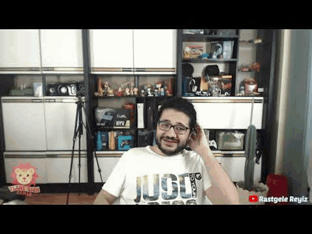 a man with glasses and a beard is sitting in front of a shelf in a room .
