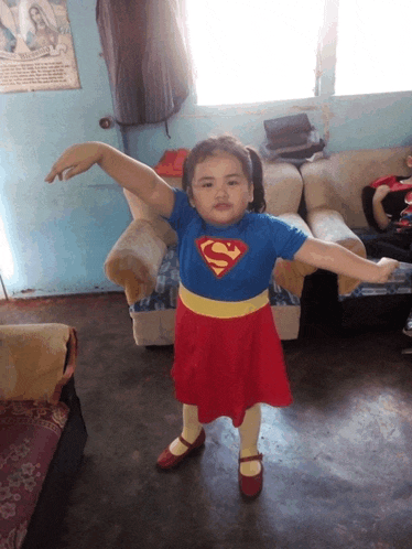 a little girl in a superman costume stands in front of a couch