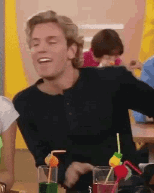 a man in a black shirt is sitting at a table with drinks and straws in front of him .