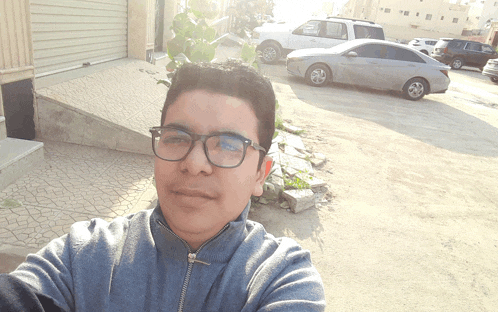 a young man wearing glasses takes a selfie in front of a parking lot
