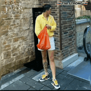 a man wearing shorts and a yellow shirt is holding a red bag in front of a sign that says t98.strong