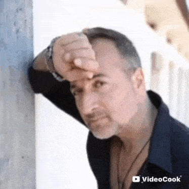 a man is leaning against a wall with his hand on his forehead and looking at the camera .