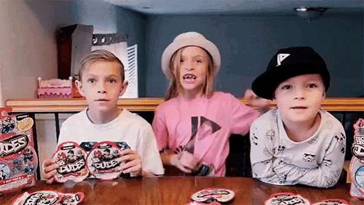 three young children are sitting at a table holding packages of dudes candy .