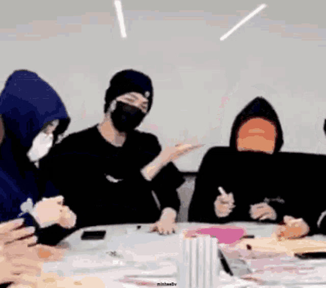 a group of people are sitting around a table wearing masks and hoodies .