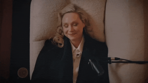a woman in a black jacket and white shirt smiles while laying on a bed