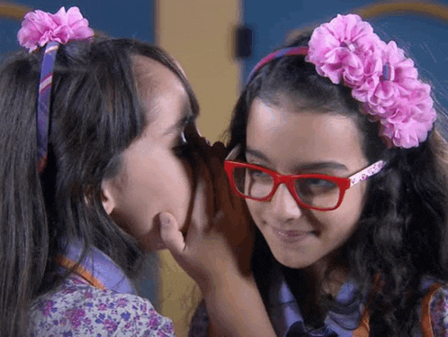 a girl with glasses is whispering into another girl 's ear .