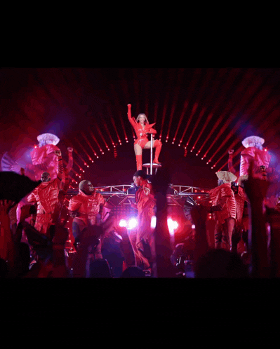 a woman in a red outfit is standing on a pole on a stage surrounded by dancers