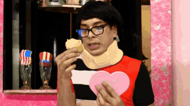 a man wearing glasses and a neck brace is eating a cupcake and holding a pink heart