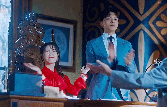 a man in a blue suit and tie is standing next to a woman in a red dress who is sitting in a chair .
